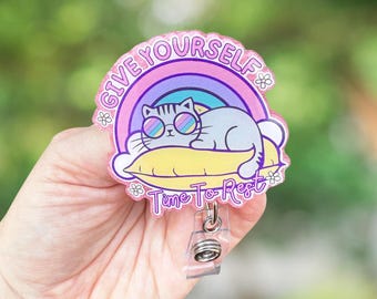 May include: A badge reel with a cartoon cat wearing sunglasses, resting on a yellow pillow under a rainbow. The text reads "GIVE YOURSELF Time To Rest". The reel has a clear plastic clip.