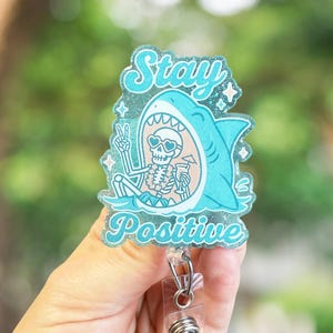 May include: A teal badge reel with the words "Stay Positive" and a cartoon shark with a skeleton inside. The skeleton is wearing sunglasses and holding a drink. The badge reel has a clear plastic clip and a retractable cord.