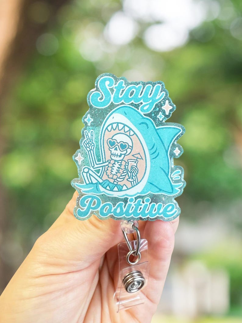 May include: A teal badge reel with the words "Stay Positive" and a cartoon shark with a skeleton inside. The skeleton is wearing sunglasses and holding a drink. The badge reel has a clear plastic clip and a retractable cord.