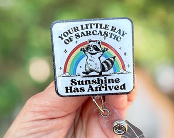 Op de afbeelding: Een witte badgehouder met een zwart-witte illustratie van een wasbeer die voor een regenboog staat. De tekst op de badgehouder luidt "Your Little Ray of Sarcastic Sunshine Has Arrived".