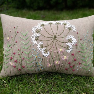 May include: A beige linen pillow with embroidered white flowers and green leaves. The pillow is on a bed of green grass.