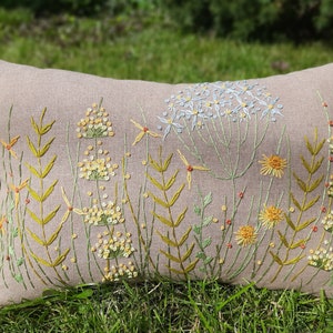 May include: A beige linen pillow with embroidered wildflowers in shades of green, yellow, and white. The pillow features a variety of flowers, including daisies, Queen Anne's Lace, and other wildflowers.