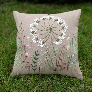 May include: A beige linen pillow with embroidered white flowers and green stems. The pillow is on a bed of green grass.