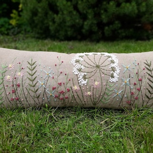 May include: A long, rectangular linen pillow with an embroidered design of a white flower with green stems and leaves. The pillow is surrounded by green grass.