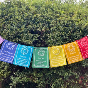 Könnte beinhalten: Ein farbenfrohes Chakra-Banner mit sieben rechteckigen Flaggen in verschiedenen Farben. Jede Flagge trägt ein Symbol und Text, die sich auf die Chakren beziehen. Das Banner hängt vor grünem Laub.