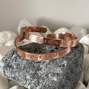 May include: Two copper bracelets with engraved messages. The top bracelet reads "Best Dad" and the bottom bracelet reads "Love You Dad".