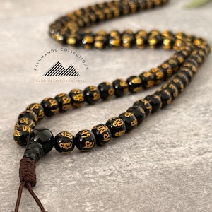 May include: A string of black prayer beads featuring gold-colored script characters on each bead. The necklace includes a larger bead and a brown cord with two dangling beads at the end. The text Kathmandu Collections Handicrafts From Nepal is visible.