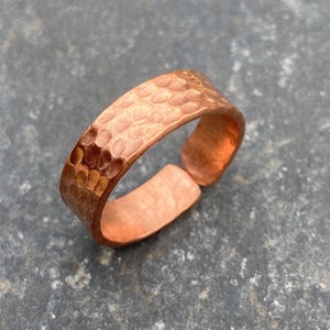 May include: A hammered copper ring with an open design. The ring has a textured surface with small, evenly spaced indentations. The copper has a warm, reddish-brown hue. The ring is set against a grey background.