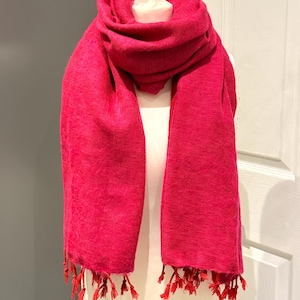 Op de afbeelding: Een levendige fuchsia sjaal met een geweven textuur is over een mannequin gedrapeerd. De sjaal heeft een zacht, vloeiend ontwerp met gefranjerde kwastjes in een bijpassende rode tint aan de uiteinden. Een opvallend modeaccessoire.