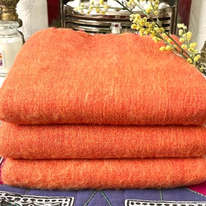 May include: A stack of three orange wool blankets folded on a patterned surface. The blankets are soft and fluffy, and they would be perfect for keeping warm on a cold day.