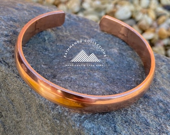 May include: A polished copper cuff bracelet with a smooth, rounded surface and an open back. The center of the bracelet features a white logo with the text KATHMANDU COLLECTIONS HANDICRAFTS FROM NEPAL and a mountain graphic.