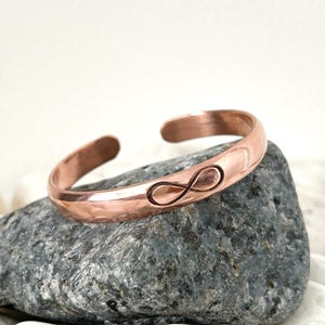 Könnte beinhalten: Ein kupferfarbenes Armband mit einem eingravierten Unendlichkeitszeichen. Das Armband hat eine gehämmerten Textur und eine rötlich-braune Farbe. Das Armband liegt auf einem grauen Stein. Der Umfang beträgt ca. 17 cm.