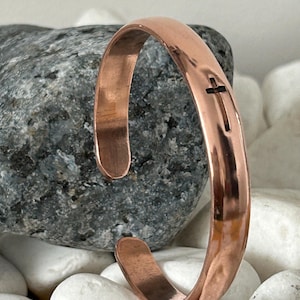 May include: A copper cuff bracelet featuring a cross cutout. The bracelet has a smooth, polished finish and an open design. The cross is centered on the front. The bracelet is displayed on a background of rocks and white stones.