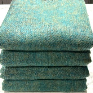 Puede incluir: Una pila de cuatro bufandas tejidas de color azul verdoso y verde. Las bufandas están dobladas y apiladas una encima de la otra.