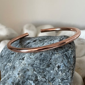 May include: A simple, polished copper bangle bracelet. The bracelet is a single, continuous piece of metal, with a smooth, rounded edge.