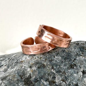May include: Two copper rings with textured, hammered designs. The rings are a warm, reddish-brown colour and are stacked on top of each other. The rings are on a grey stone surface.