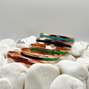 May include: A stack of copper cuff bracelets featuring inlaid stone strips in turquoise, dark blue, green, and red. Each bracelet has a smooth metallic finish and a central channel filled with colorful stone segments. The logo Kathmandu Collections is visible.