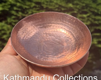 May include: A small, round, hammered copper bowl with a metallic finish and a reflective surface. The text Kathmandu Collections is printed in white at the bottom of the image, overlaying the bowl and the background.