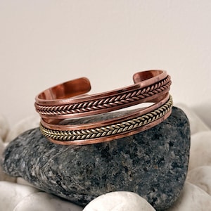 Op de afbeelding: Koperen manchetarmband met een gevlochten ontwerp. De armband heeft een warme koperkleur met een contrasterende goudkleurige band. De manchet wordt getoond op een grijze steen, omringd door witte kiezelstenen.