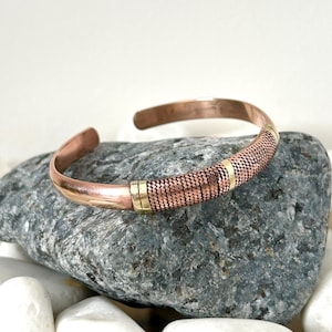 May include: A copper bracelet with a braided design and gold accents. The bracelet is open and adjustable.