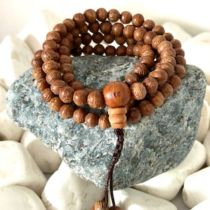 Può includere: Un braccialetto di perline mala in legno marrone con un nappa di corda marrone. Il braccialetto è fatto di piccole perline rotonde con motivi intricati. Il braccialetto è appoggiato su una roccia grigia.