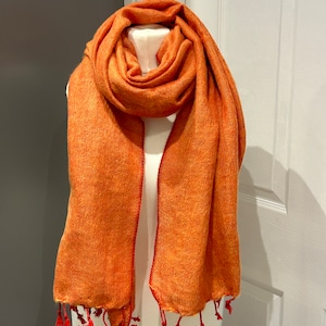 Puede incluir: Una suave bufanda naranja con flecos rojos. La bufanda está hecha de un material ligero y es perfecta para capas.