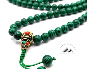 May include: A long strand of green malachite beads featuring a central brass spacer with inlaid red and turquoise stones. The necklace ends in a green cord with two dangling malachite beads. The text Kathmandu Collections is visible.