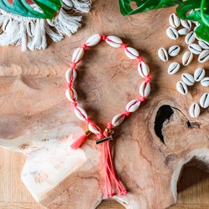 Peut inclure: Un collier de coquillages rose et blanc avec un pompon rose. Le collier est composé de petites coquilles blanches enfilées sur un fil rose. Le pompon est en fil rose et est attaché au collier par un fermoir doré.