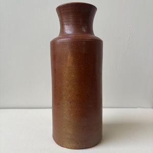 Könnte beinhalten: Eine braune Steinzeugvase mit zylindrischem Körper und leicht ausgestelltem Hals. Die Vase hat eine glatte, glasierte Oberfläche mit Farbvariationen, die von einem tiefen Braun bis zu einem helleren, goldenen Farbton in der Nähe des Bodens reichen. Die Vase ist etwa 25 cm hoch.
