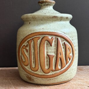 Pode incluir: Um açucareiro de cerâmica bege salpicado com tampa a condizer. A parte frontal apresenta um design oval em relevo com a palavra SUGAR gravada em letras maiúsculas grandes de cor castanho-escuro. O acabamento tem um aspeto texturado.