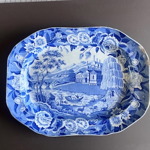 May include: Blue and white ceramic serving platter with a floral border and a scenic landscape design. The landscape features a river, bridge, buildings, and trees. The platter has an octagonal shape.