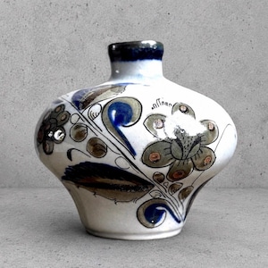 May include: A white ceramic vase with a wide, rounded body and a narrow neck. The surface features hand-painted floral motifs in shades of dark blue, muted green, and brown, with a dark blue rim around the opening of the vase.