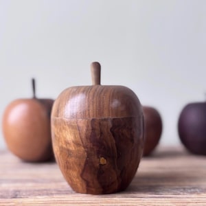 May include: A wooden apple-shaped container with a stem-like handle. The container is made of dark brown wood with visible grain patterns. Other wooden apple-shaped objects are in the background, with varying shades of brown and purple.