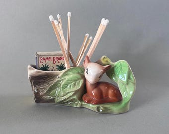 May include: A ceramic match holder shaped like a tree stump with a deer figurine sitting on a green leaf. The holder has a compartment for matches and a box of "Camel Brand" matches is inside.