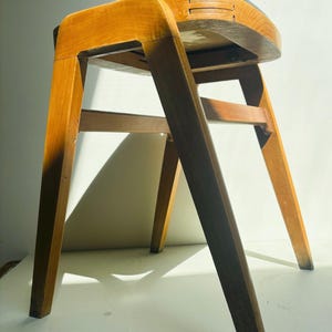 May include: A wooden stool with four angled legs and a curved seat. The light-toned wood frame features visible joinery details on the seat edges. The seat is topped with a smooth, light grey surface, and the structure includes horizontal support bars.