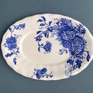 Op de afbeelding: Ovale witte keramische schaal met een blauw bloemmotief. De schaal heeft een geschulpte rand en een gedetailleerd patroon van blauwe bloemen en bladeren. Het ontwerp is geconcentreerd aan één kant van de schaal.