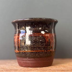 Op de afbeelding: Een kleine keramische beker met een uitlopende rand, voorzien van een donkerbruin gespikkeld glazuur op het bovenste gedeelte en een matte terracotta basis. Een horizontale oranje band omcirkelt het midden met verticale oranje druppels.