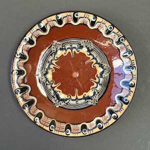 May include: A round ceramic plate with a glossy terracotta-coloured centre and a decorative border. The design features intricate, marbled patterns in cream, dark blue, and black, with small blue dots encircling the outer rim of the plate.