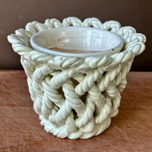 May include: A cream-coloured ceramic planter featuring a decorative exterior layer of sculpted, intertwined rope-like patterns. The vessel has a smooth, circular inner rim and a wide, open top, showcasing a glossy finish over the textured design.