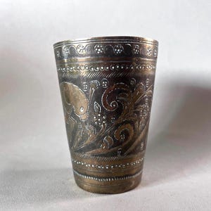 Può includere: Una piccola tazza di metallo argentata con intricati disegni floreali e geometrici incisi sulla superficie. La tazza ha un fondo arrotondato e un bordo leggermente svasato.