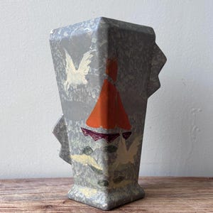 May include: Gray, geometric vase with a textured surface and painted details. The vase features a design with an orange shape, white birds, and other colorful accents. The vase has a rectangular shape with a flared top and a small base.