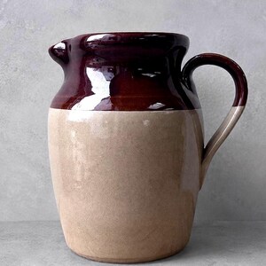 May include: A ceramic pitcher featuring a two-tone design with a glossy dark brown glaze on the upper portion and a matte beige finish on the lower body. The vessel includes a curved handle on the right side and a small pouring spout at the top.