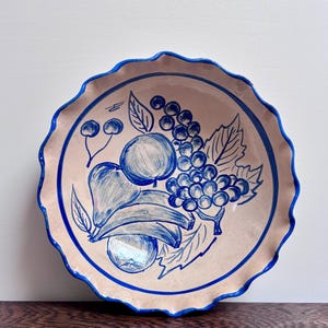 May include: A ceramic plate with a scalloped edge, featuring a hand-painted design of fruit in shades of blue on a beige background. The fruit includes grapes, apples, and bananas. The rim and the fruit outlines are painted in a vibrant blue.