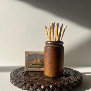 May include: A brown ceramic match holder filled with matches, next to a vintage matchbox with a ship illustration. The items rest on a dark wooden, intricately carved, round tray. The scene is lit by natural light.