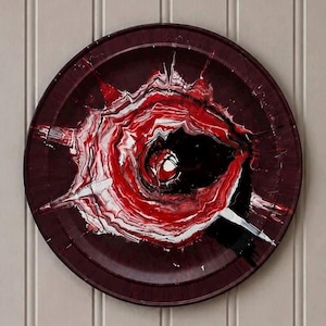 May include: A circular decorative plate featuring an abstract fluid paint design. The surface displays a swirling pattern of deep burgundy, bright red, stark white, and solid black colours, creating a dynamic, starburst-like effect across the centre.