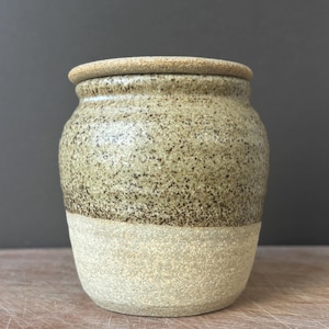 Puede incluir: Un tarro de cerámica con tapa que presenta un esmalte moteado en tonos beige y marrón en la parte superior y un acabado mate texturizado de color crema en la base. El recipiente tiene un cuerpo redondeado, un cuello corto y una tapa plana.