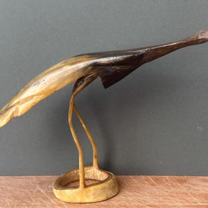 May include: A sculpture of a bird in flight, crafted from polished horn material. The piece features a gradient of colours ranging from light cream and tan at the tail to a deep, dark brown at the head and wings, supported by a circular base.