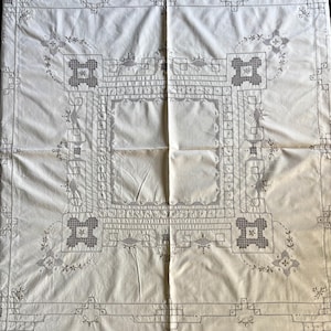 May include: A square off-white tablecloth featuring intricate dark gray embroidery. The design includes a central square border, geometric corner motifs, and delicate floral accents arranged in a symmetrical pattern across the fabric surface.