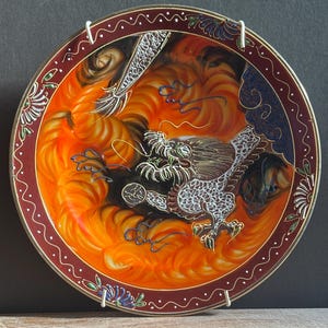 May include: A round decorative plate featuring a central illustration of a dragon with white scales and black details, surrounded by swirling orange and yellow flames. The border is dark red with white dots and blue floral accents.