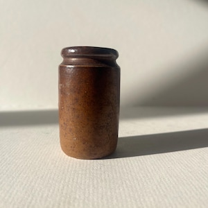 Puede incluir: Un pequeño tarro cilíndrico de gres marrón con una parte superior redondeada y una superficie ligeramente texturizada. El tarro es de un color marrón cálido y terroso y parece ser vintage o antiguo. El tarro probablemente se utiliza para el almacenamiento.
